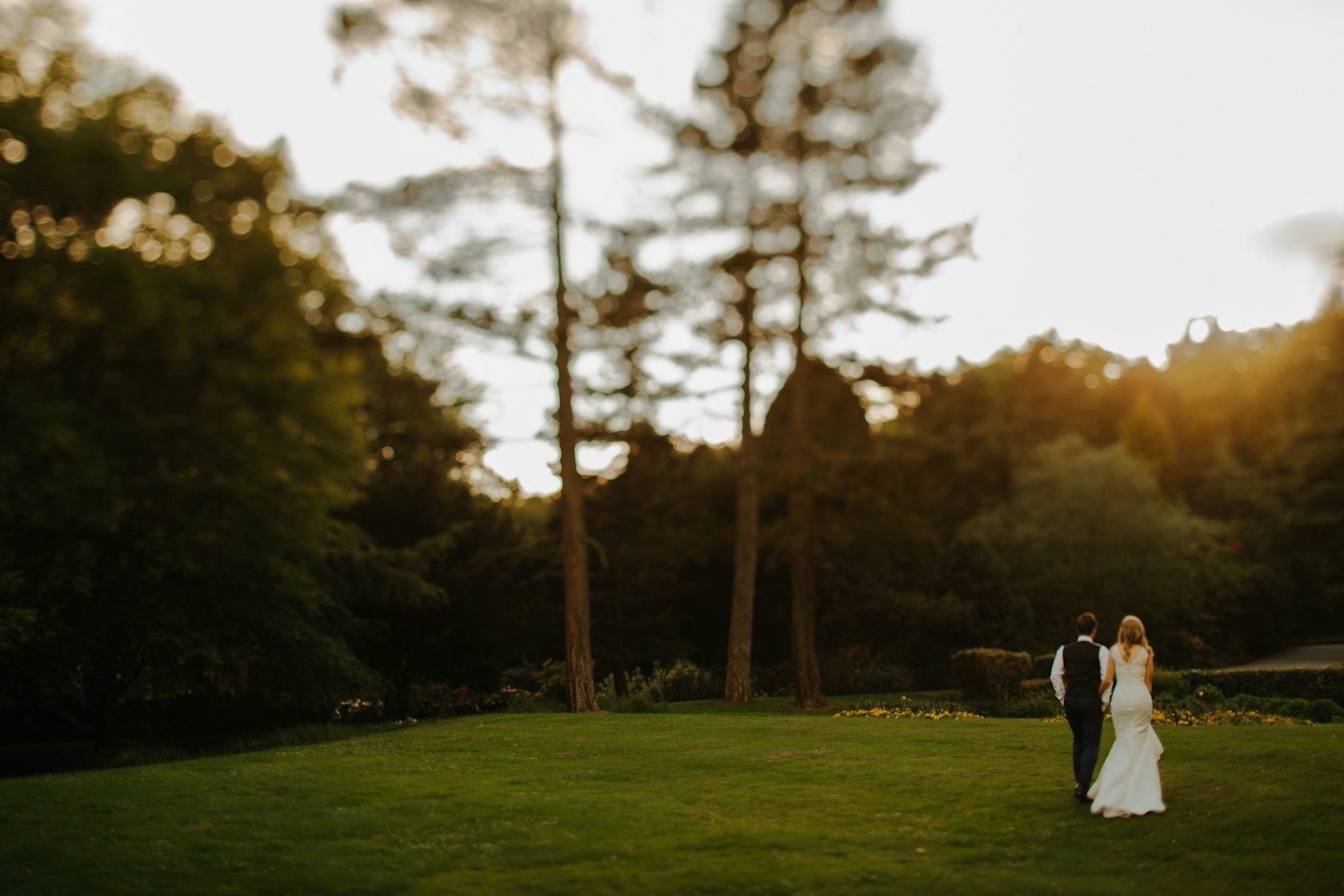 Bride and groom walk across a lawn at sunset.