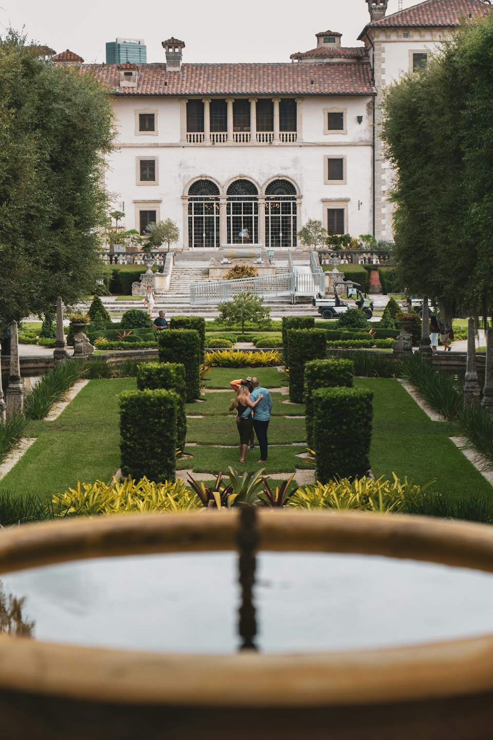 A couple embraces in a formal garden before a white mansion.