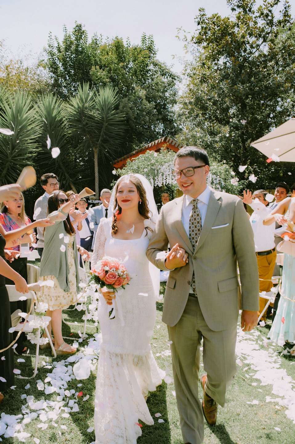 A bride and groom walk through petals in a garden.