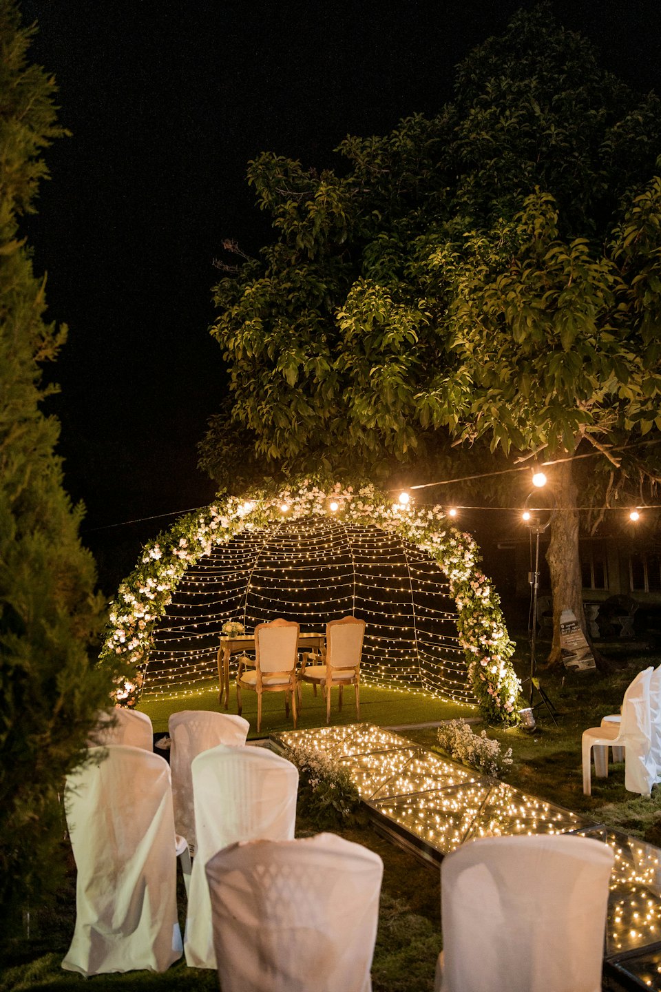 Outdoor event venue archway with lights and flowers at night.